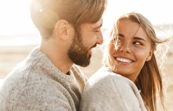 A couple wearing sweaters smiles at each other while sitting outdoors on a sunny day.